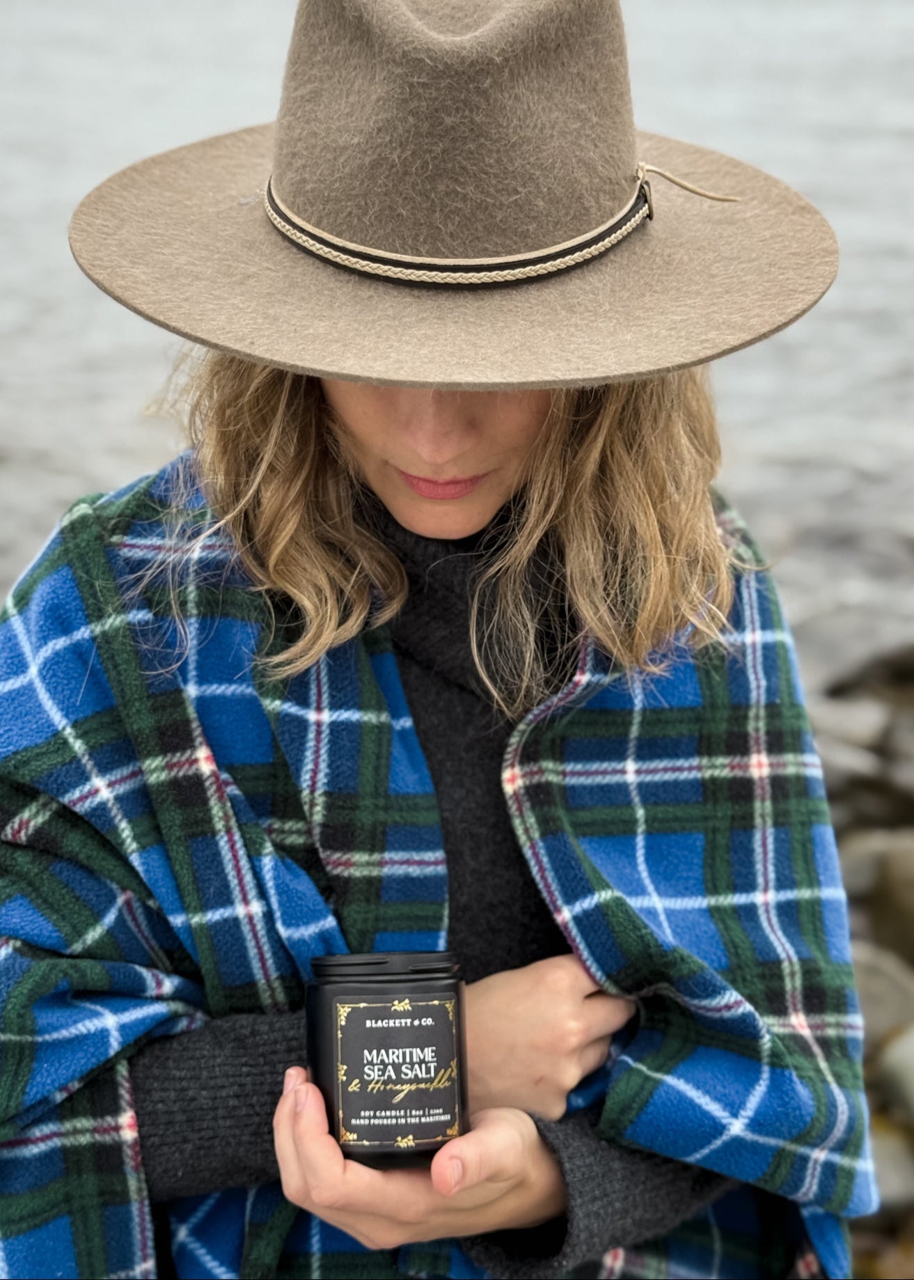 Owner of Blackett & Co. wearing a plaid Nova Scotia tartan blanket and hat, holding a black jar of 'Maritime Sea Salt & Honeysuckle' scented natural soy candle container by the ocean.