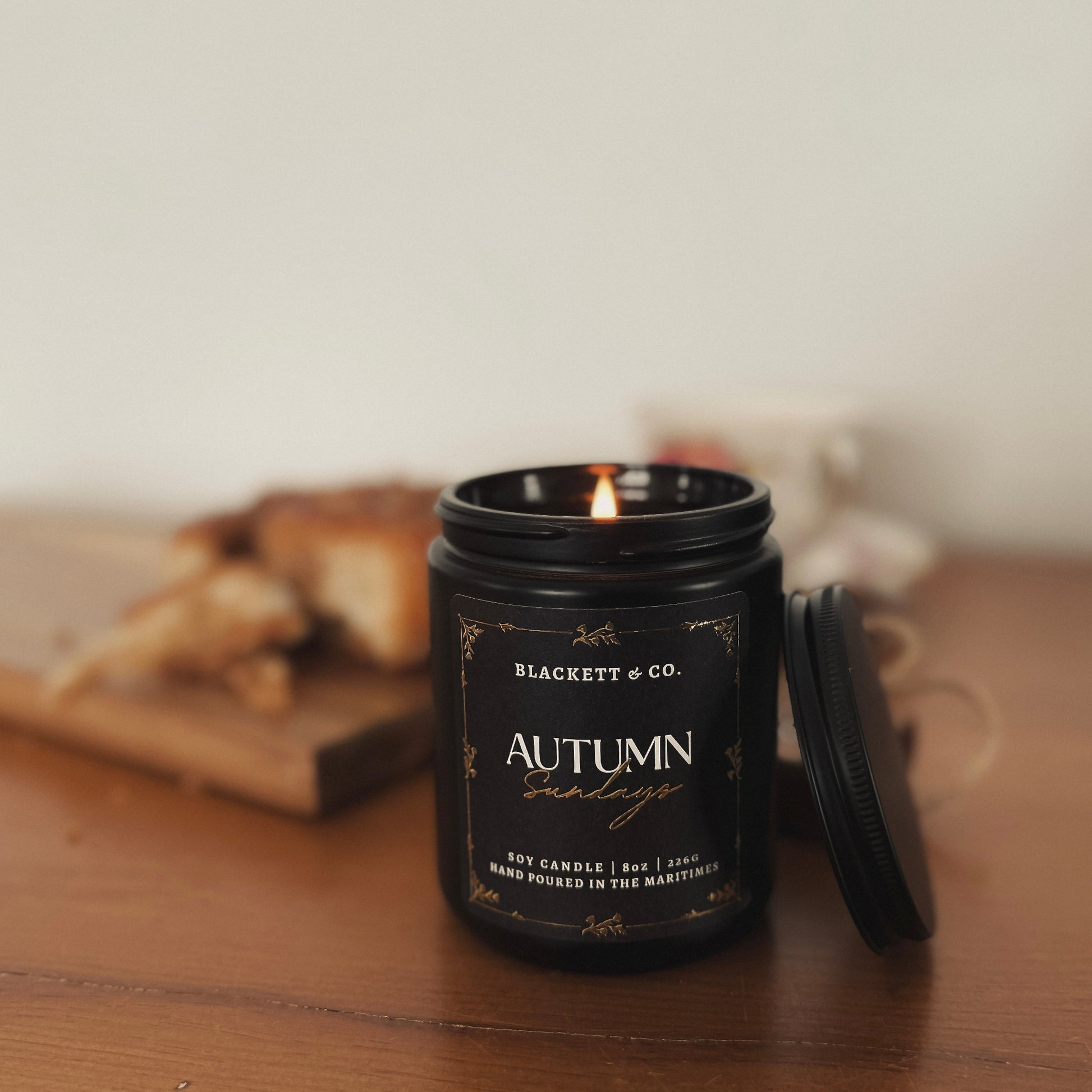 Autumn Sundays scented natural soy candle on a wooden table with pastries and a china tea cup and saucer in the background