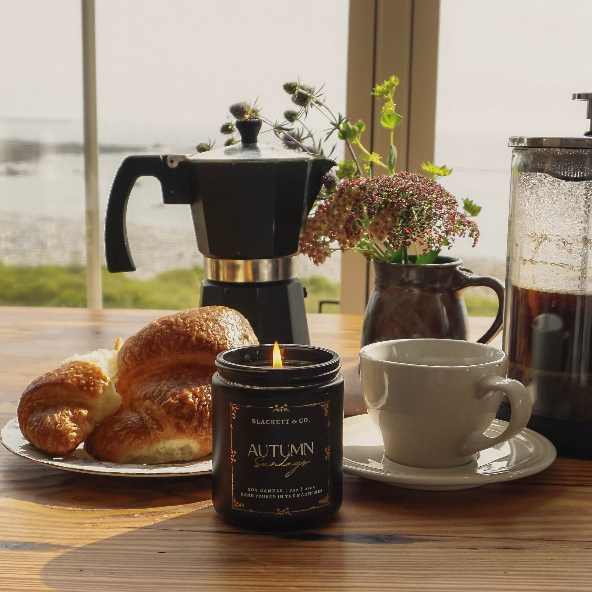 Autumn Sundays scented natural soy candle on a wooden table with pastries, a mug and saucer, and coffee pot in front of an ocean window view