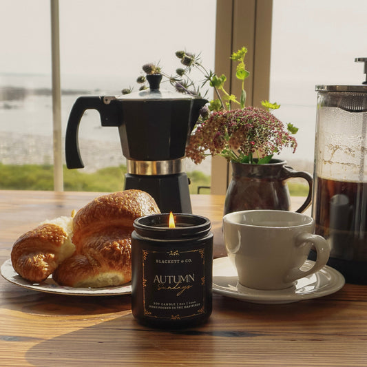 Autumn Sundays scented natural soy candle on a wooden table with pastries, a mug and saucer, and coffee pot in front of an ocean window view