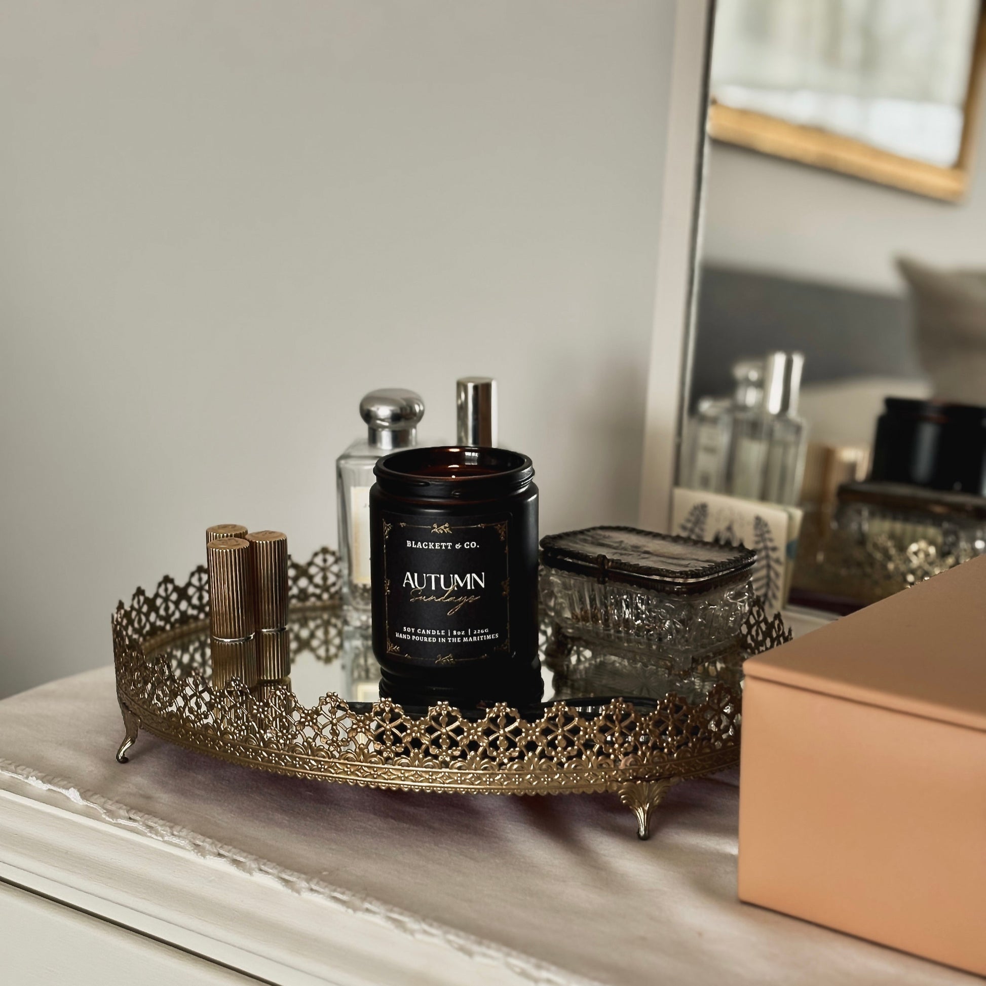 An Autumn Sundays scented natural soy candle by Blackett & Co. sit on a mirror gold trim tray surrounded by perfumes and cosmetics.