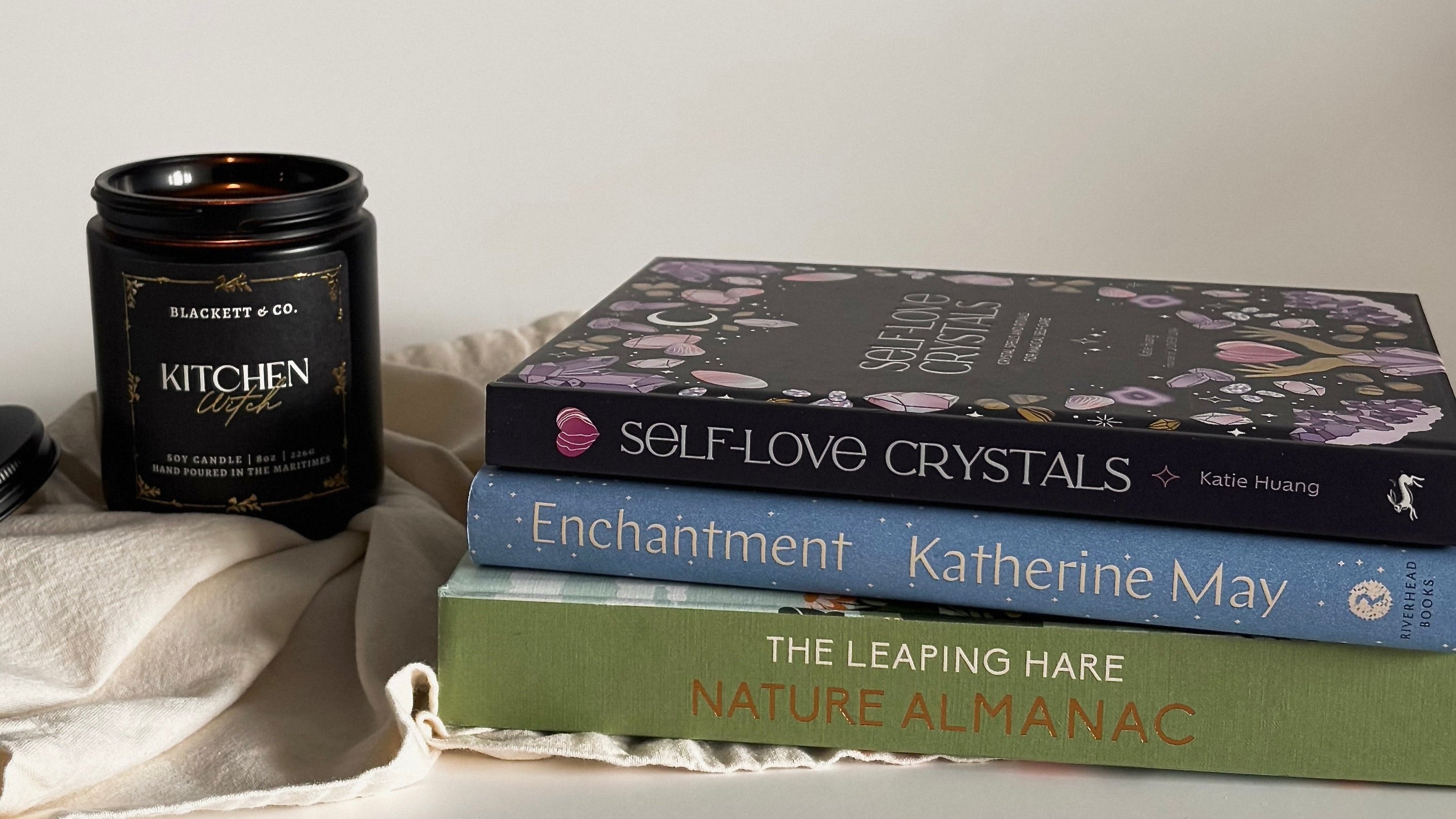 Stack of books including 'Self-Love Crystals', 'Enchantment', and ' Nature Almanac', with a Kitchen Witch scented natural candle on a white surface.