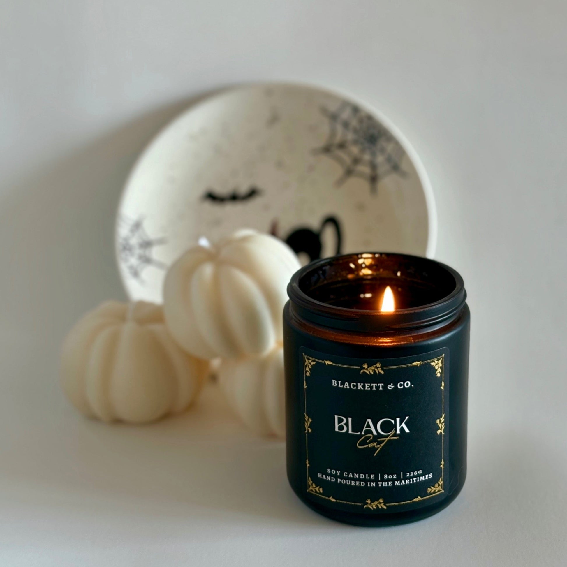 Black Cat soy candle by Blackett & Co. on a white surface with pumpkin-shaped candles and a Hallowe'en decorative plate in the background.