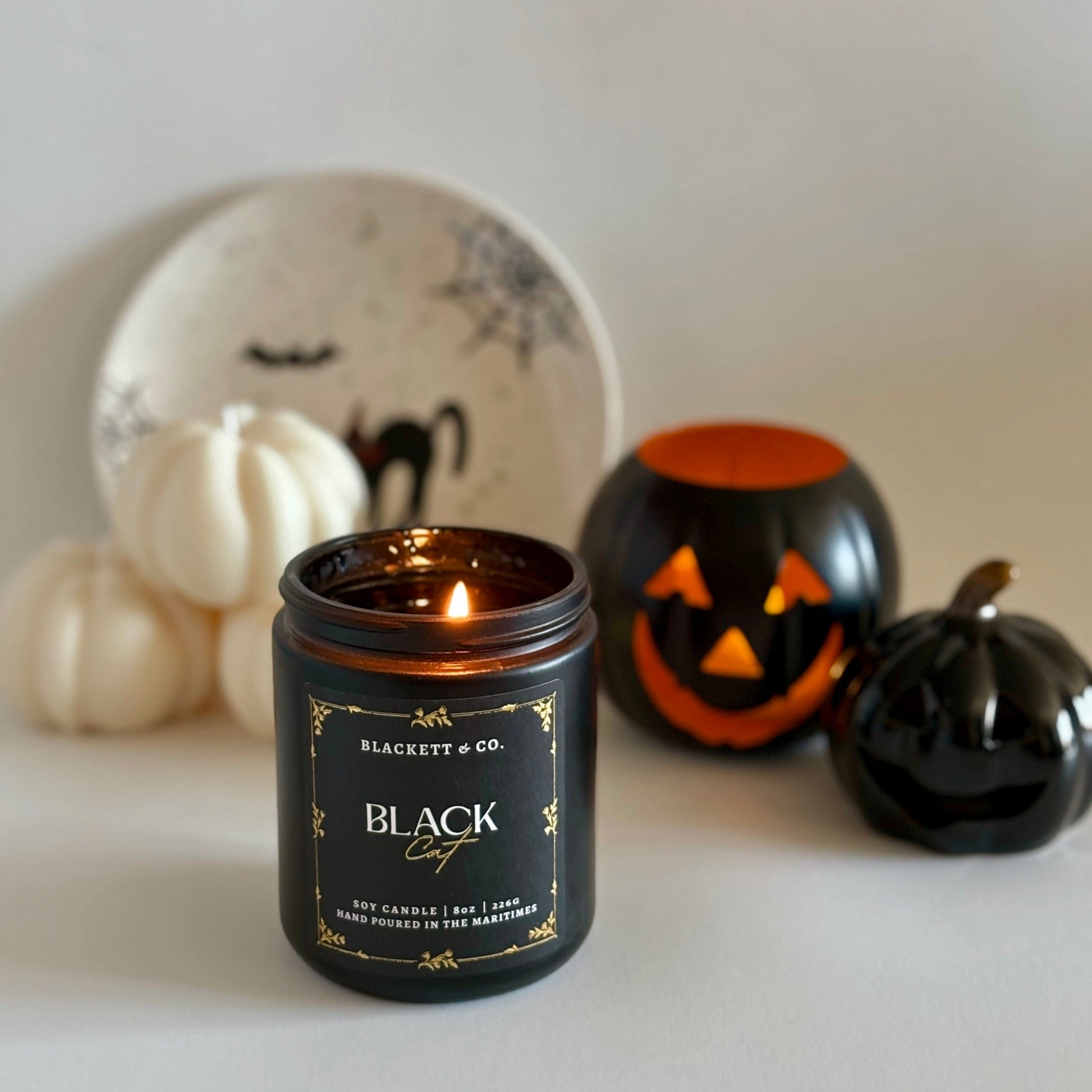 Black Cat soy candle by Blackett & Co. on a white surface with pumpkin-shaped candles and a Hallowe'en decorative plate in the background.