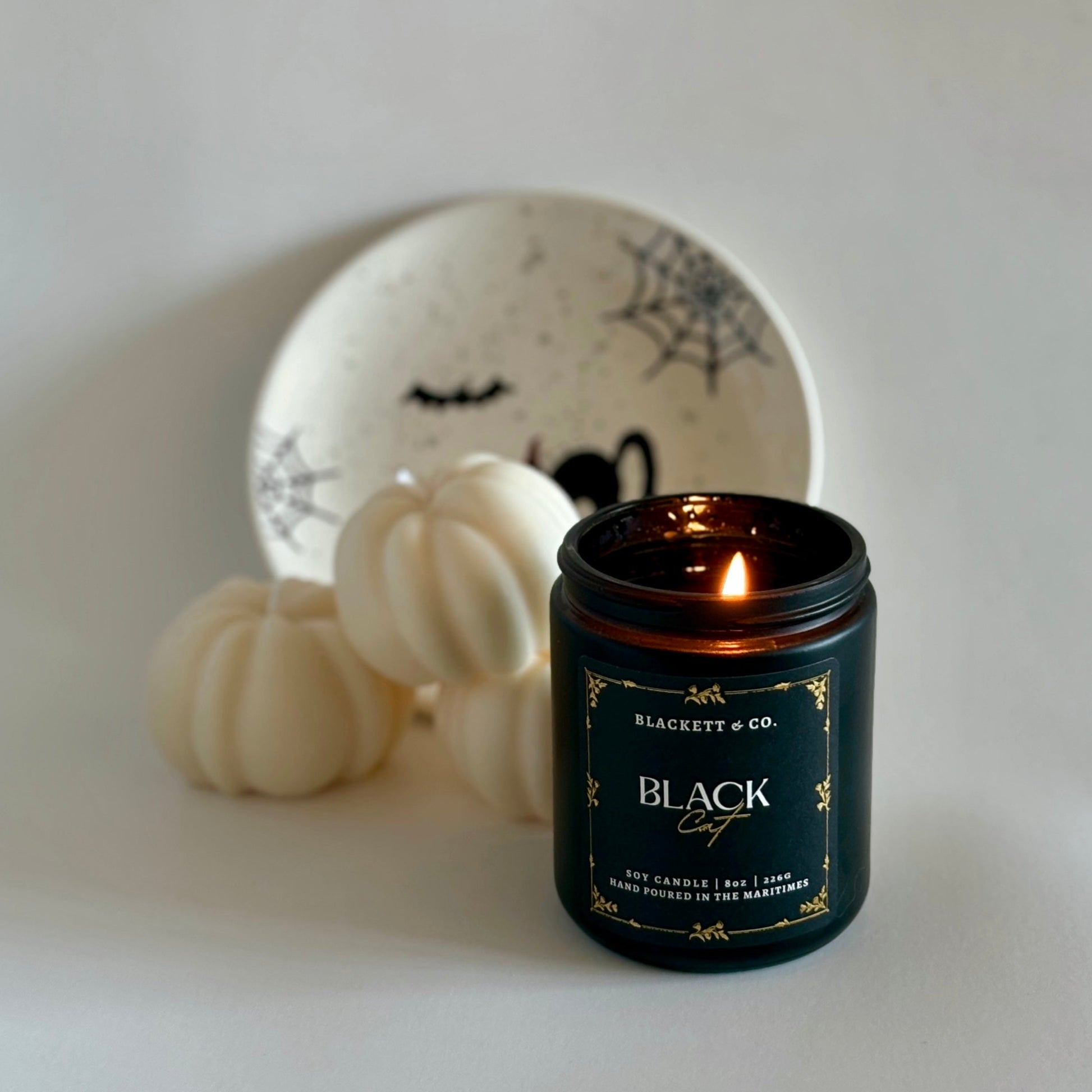 Black Cat soy candle by Blackett & Co. on a white surface with pumpkin-shaped candles and a Hallowe'en decorative plate in the background.