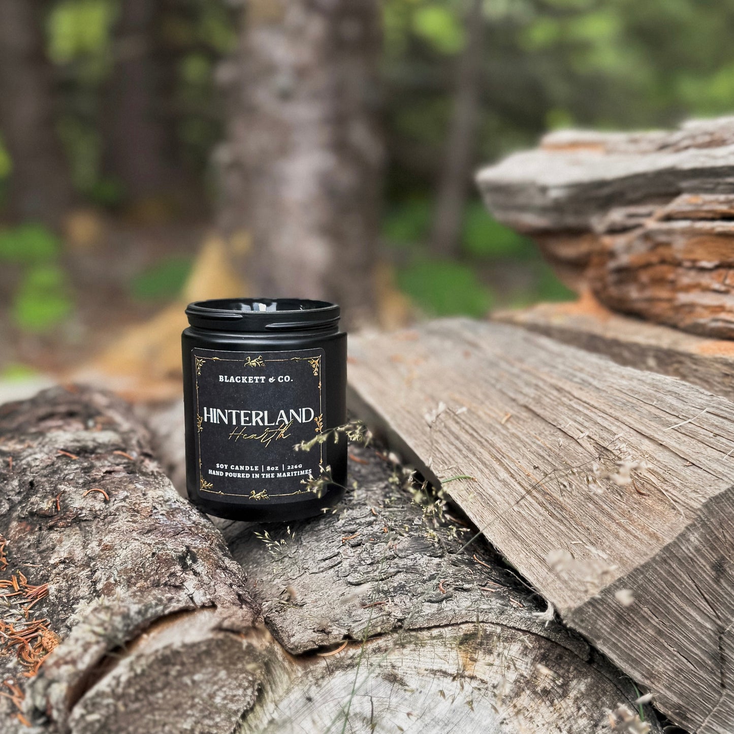 Hinterland Hearth scented soy candle in a black jar by Blackett & Co. sitting on a stack of wooden logs