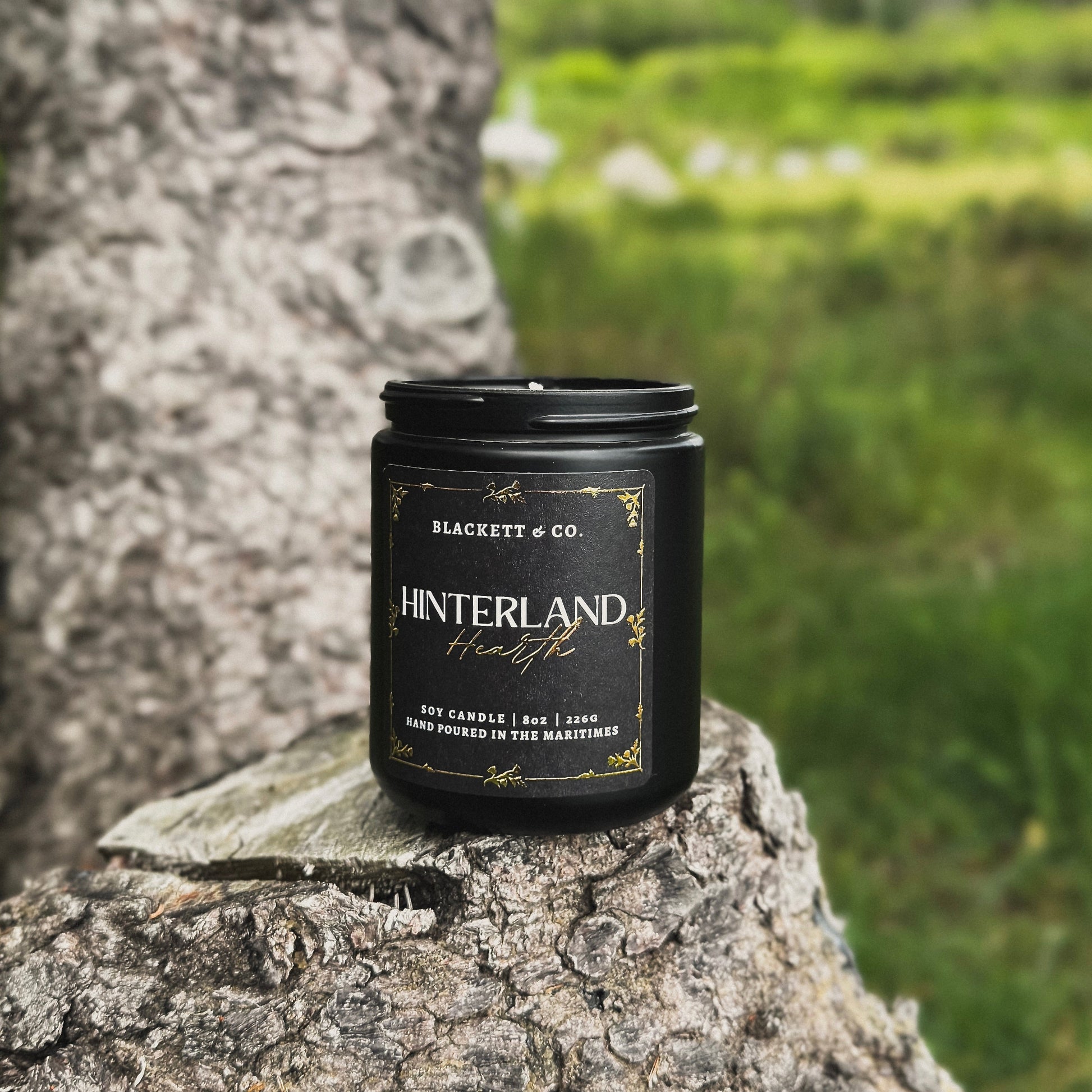 Hinterland Hearth scented natural soy candle on a tree stump with a grassy background