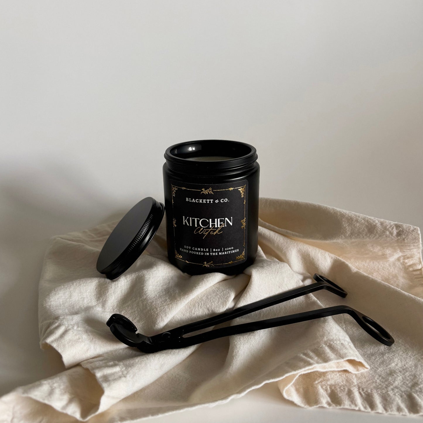 Kitchen Witch scented natural soy candle Black candle jar on a white cloth with a neutral background and a matte black wick trimmer