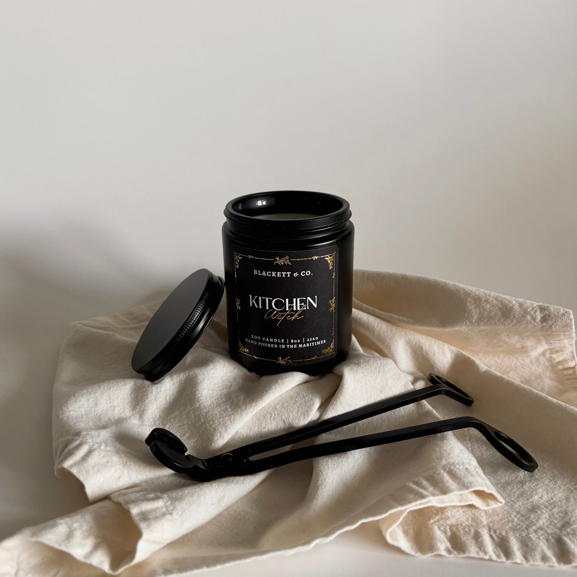 Kitchen Witch scented natural soy candle Black candle jar on a white cloth with a neutral background and a matte black wick trimmer