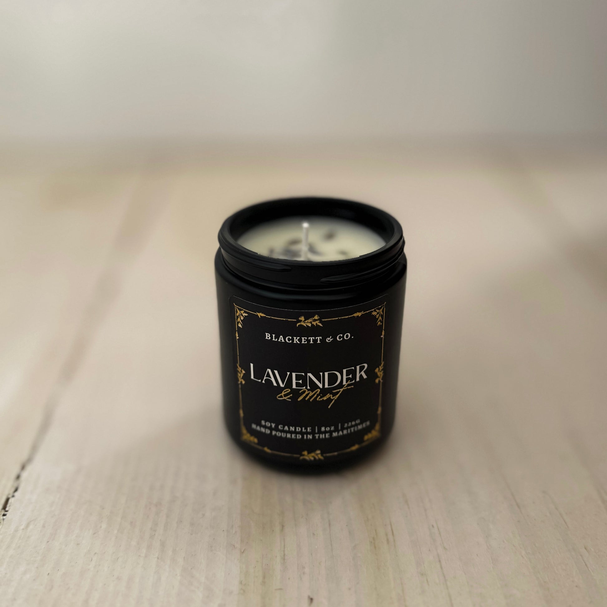 Lavender & Mint scented natural soy candle in a black candle jar by Blackett & Co. on a light wooden surface