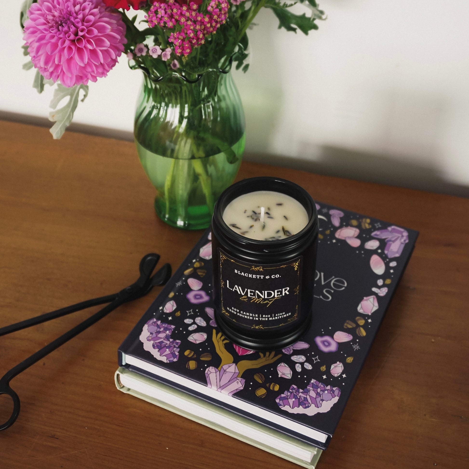 Lavender & Mint scented natural soy candle on a stack of books with a bouquet of flowers and a matte black wick trimmer