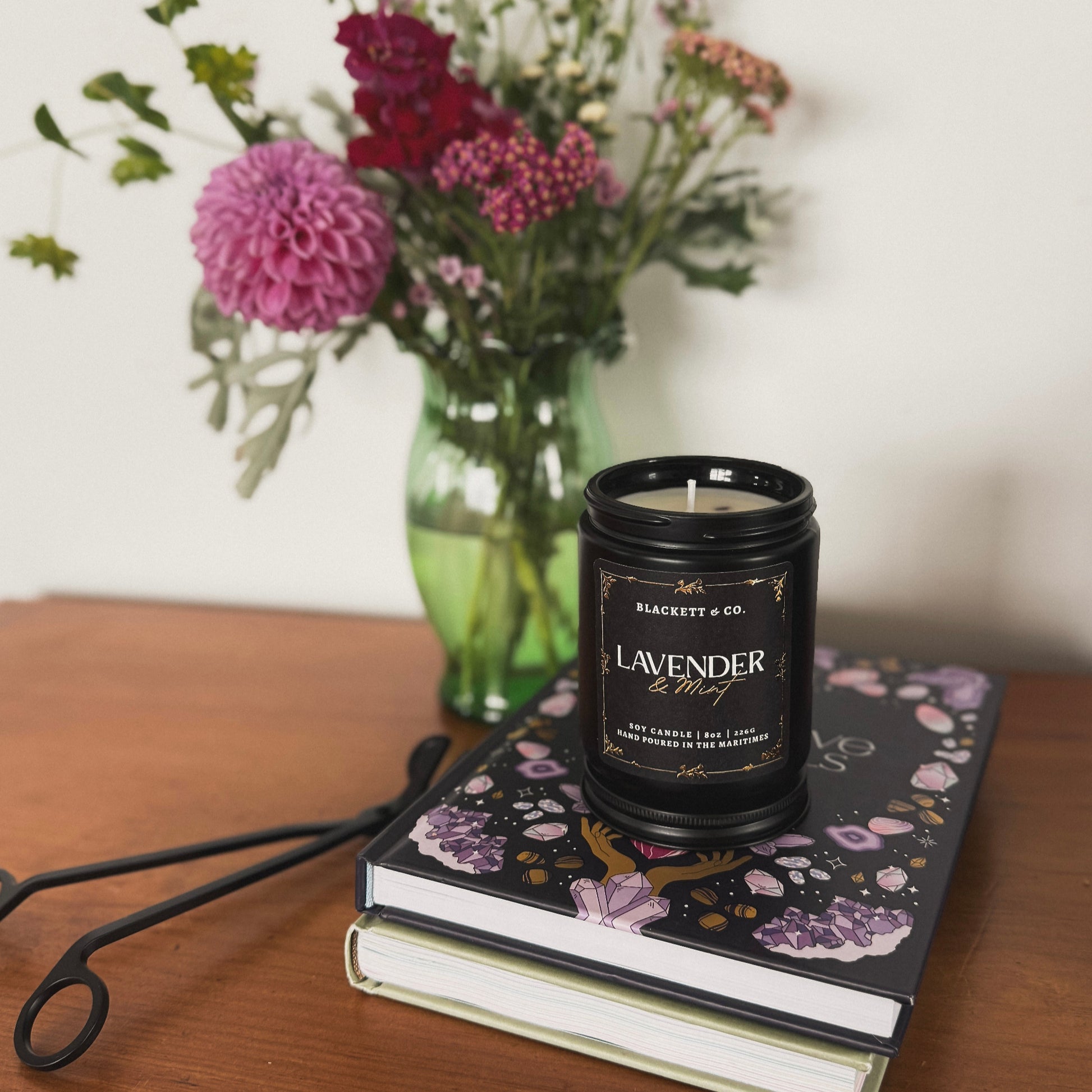 Lavender & Mint scented natural soy candle in a black jar by Blackett & Co. on a stack of books with flowers in the background