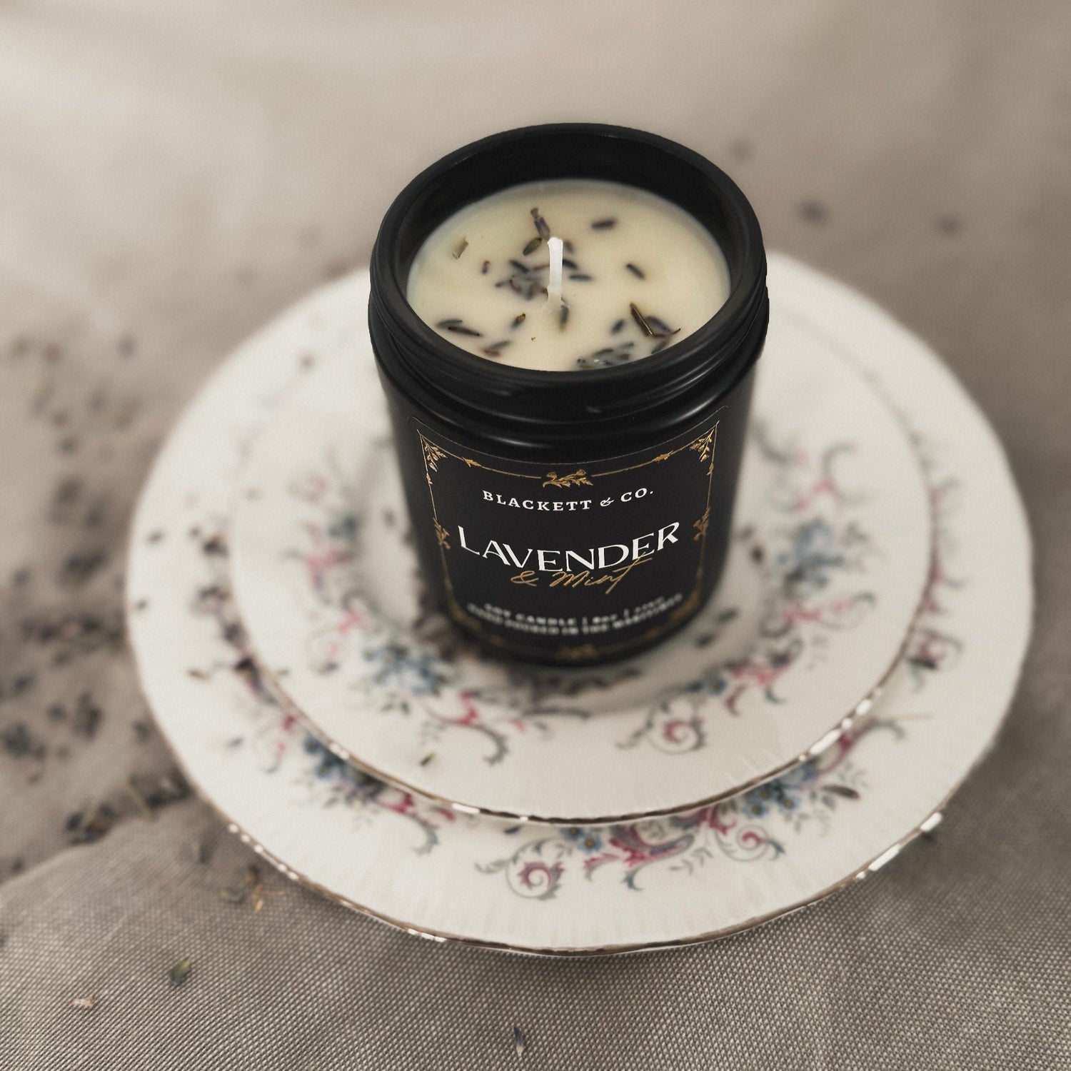 Lavender & Mint scented natural soy candle sitting a small stack of floral china dishes with Nova Scotian grown lavender buds
