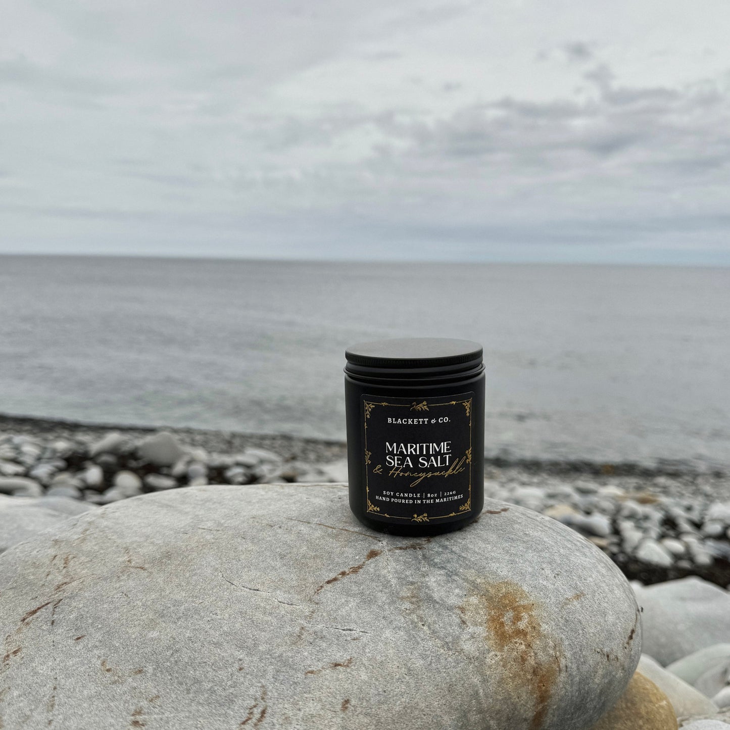 Maritime Sea Salt & Honeysuckle scented natural soy candle on a pebble beach with ocean and sky in the background