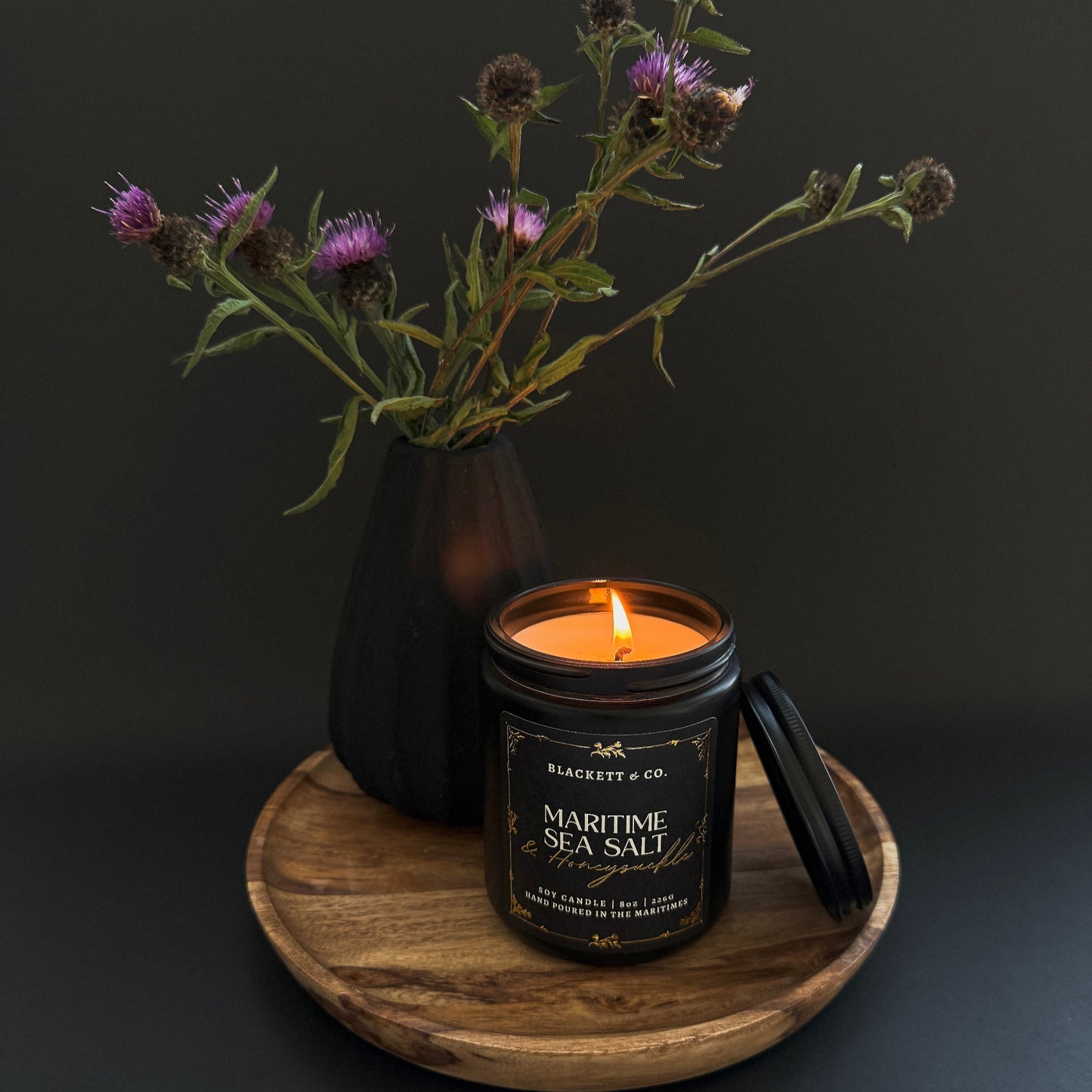 Maritime Sea Salt & Honeysuckle scented natural soy candle in a black candle by Blackett & Co. on a wooden plate and a black vase with thistles