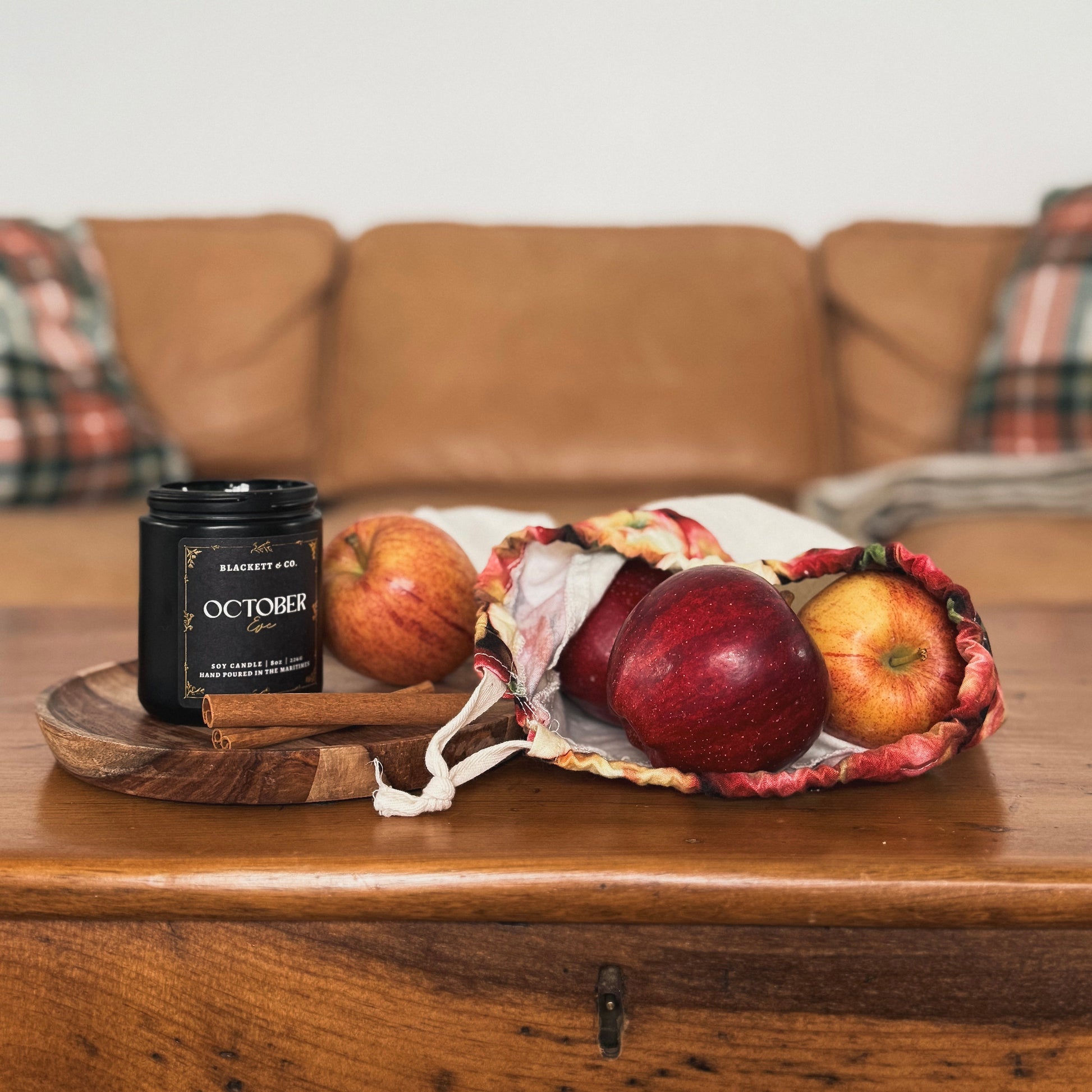 October Eve scented natural soy candle in a black jar by Blackett & Co. next to apples on a wooden surface with a couch in the background