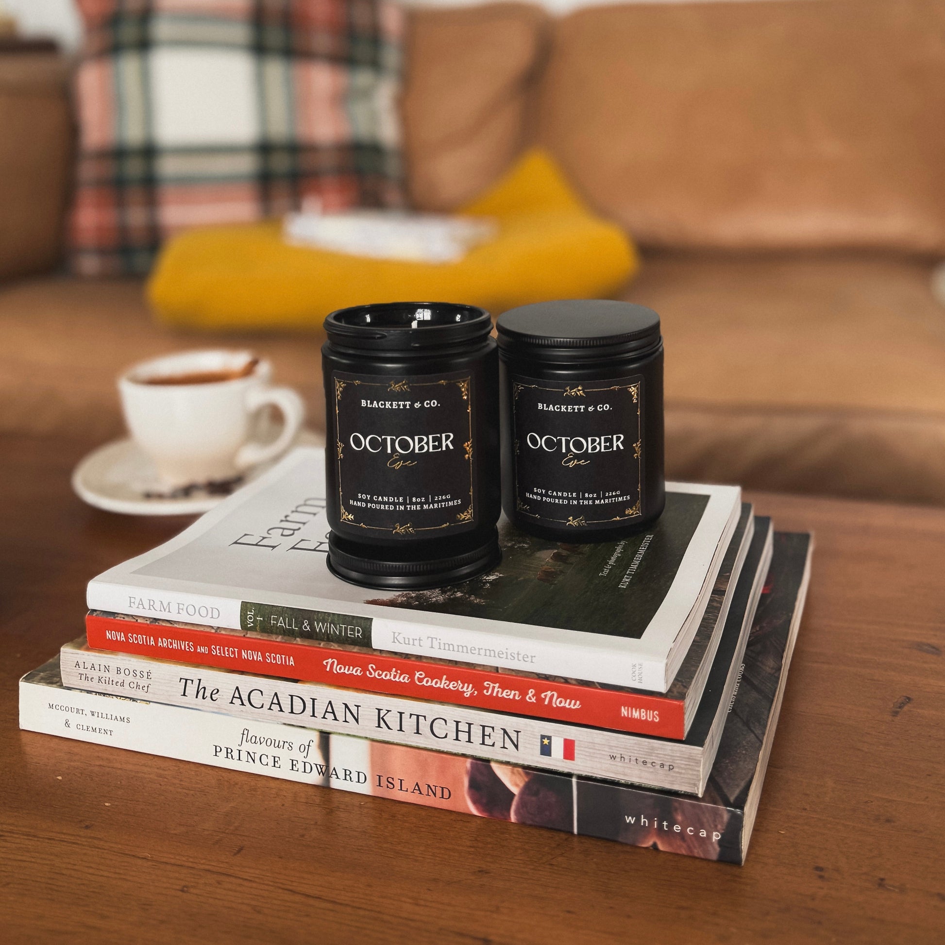 October Eve scented natural soy candle in a black jar by Blackett & Co. on a stack of books with a mug of apple cider in the background