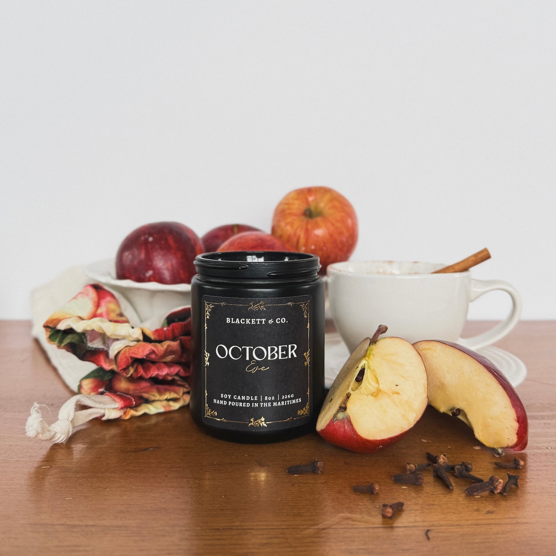 October Eve scented natural soy candle in a black jar by Blackett & Co. with apples and a mug of apply cider on a wooden surface