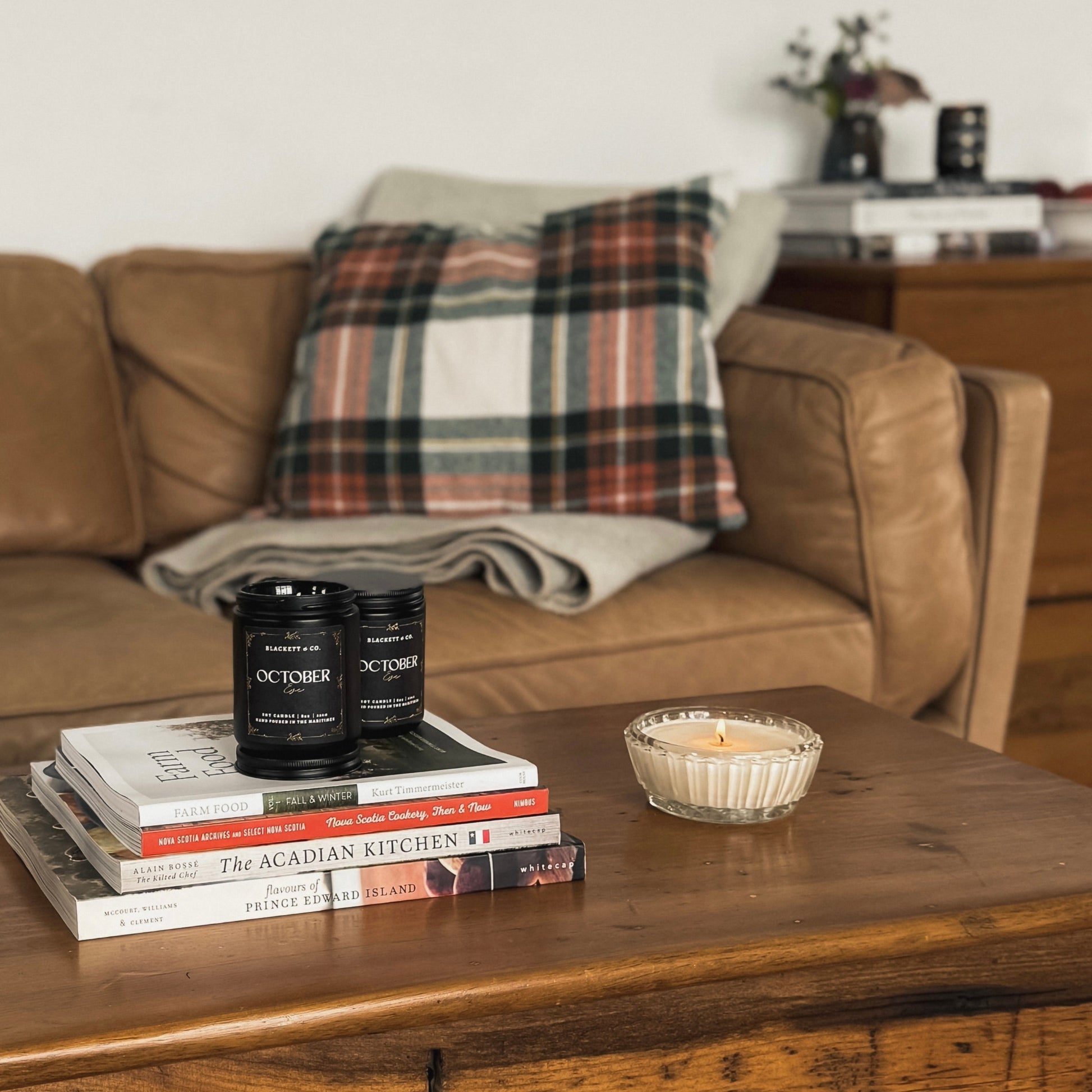 October Eve scented natural soy candles on a stack of cook books in front of a brown leather sofa with plaid pillow and wooden coffee table with books and candle in the background
