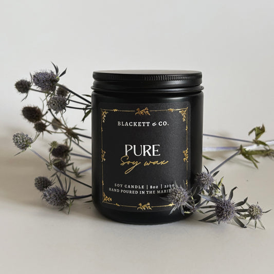 Pure unscented soy candle in a black jar by Blackett & Co. against a light background and thistles