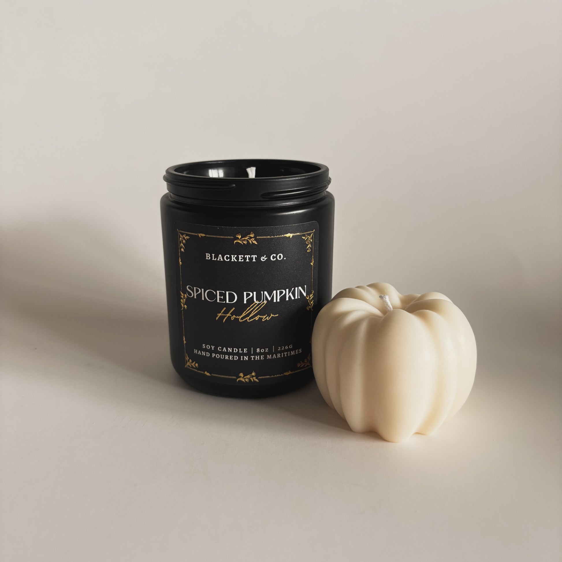Spiced Pumpkin Hollow scented soy candle in a black jar by Blackett & Co. with a small white soy wax pumpkin on a light background