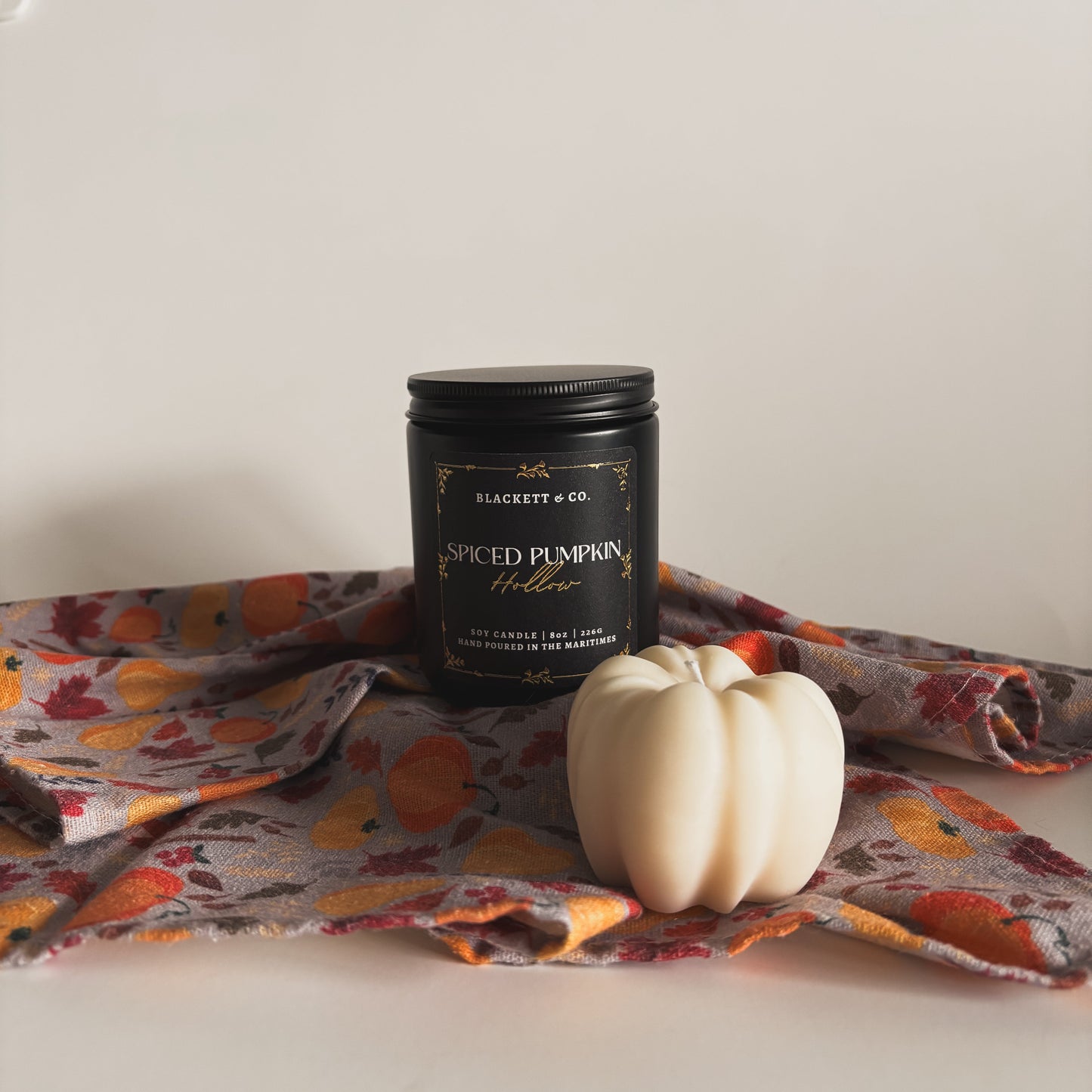 Spiced Pumpkin Hollow scented soy candle in a black jar by Blackett & Co. with a small white soy wax pumpkin candle on autumnal pattern linen against a light background
