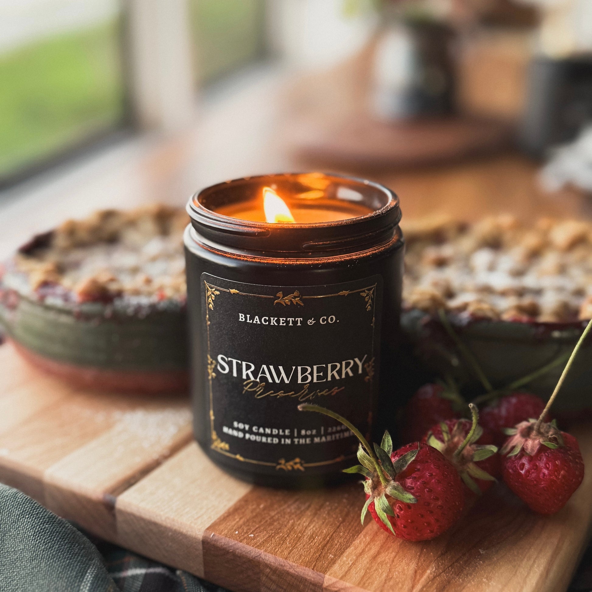 Strawberry Preserves scented natural soy candle in a black jar by Blackett & Co. on a wooden cutting board with strawberry crips and berries