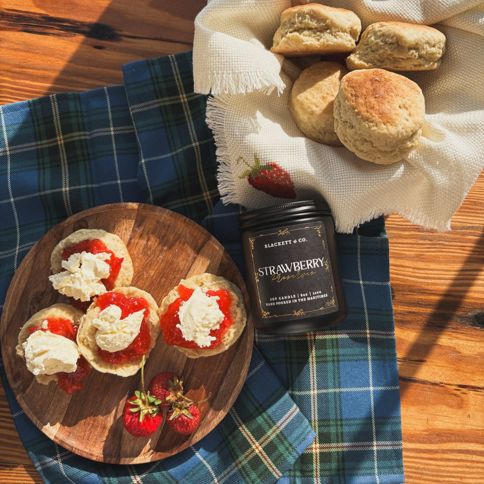 Strawberry Preserves scented natural soy candle surrounded by biscuits with strawberry jam and cream on a wooden board on a Nova Scotia tartan cloth