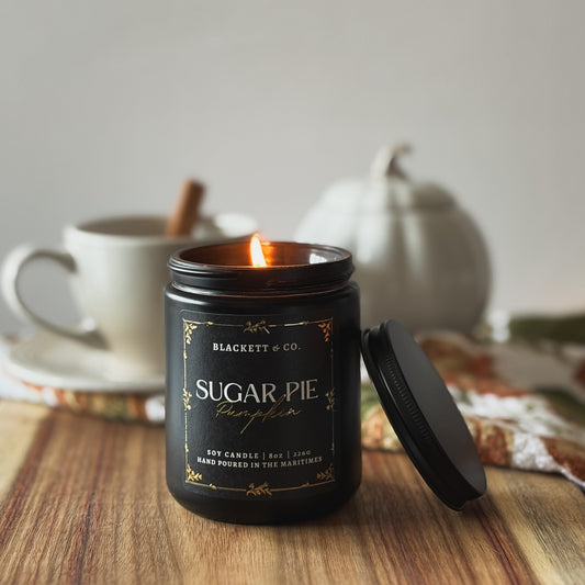 Sugar Pie Pumpkin scented natural soy candle by Blackett & Co. on a wooden surface with a white coffee cup and white ceramic pumpkin blurred background.