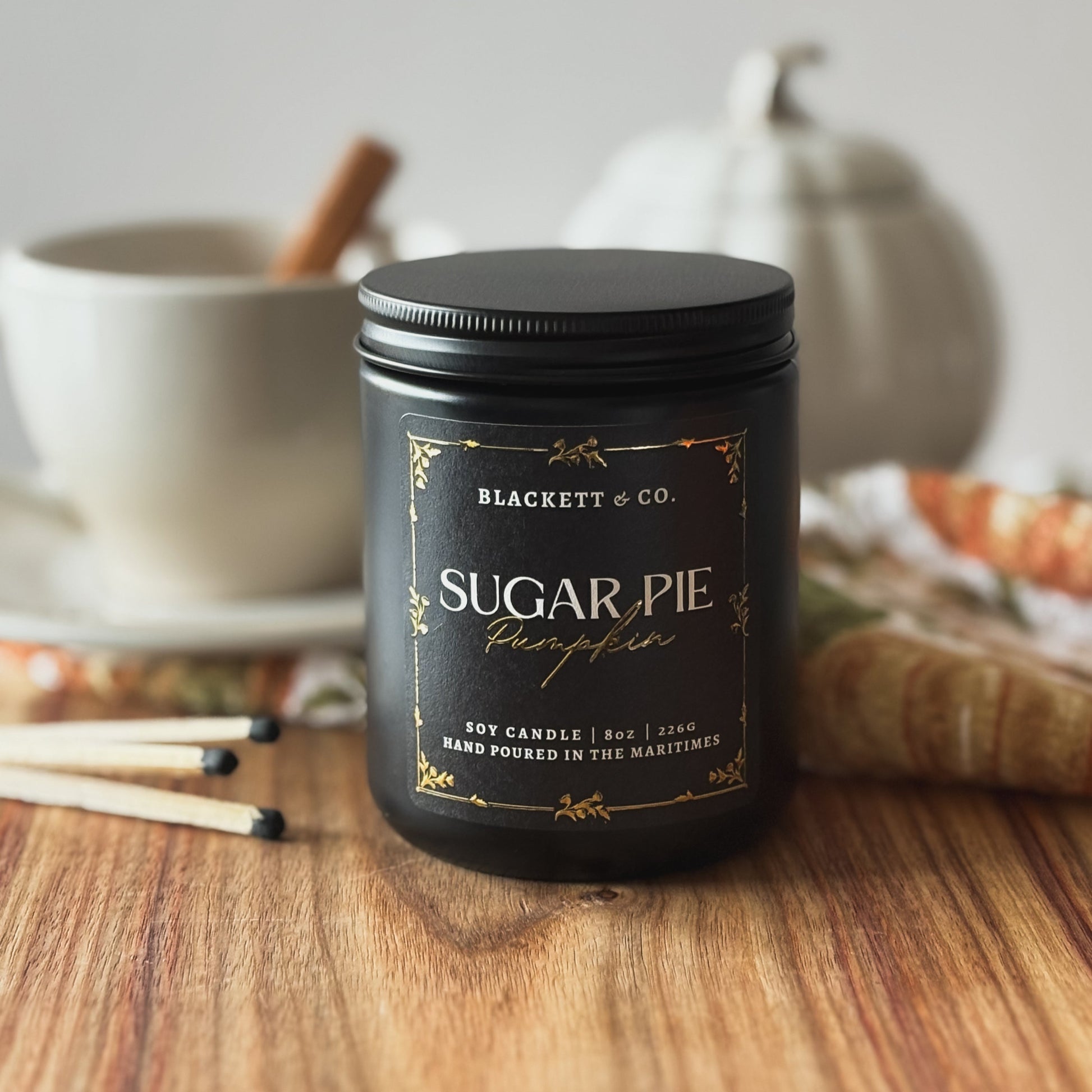 Sugar Pie Pumpkin scented natural soy candle by Blackett & Co. on a wooden surface with a white coffee cup and white ceramic pumpkin blurred background.