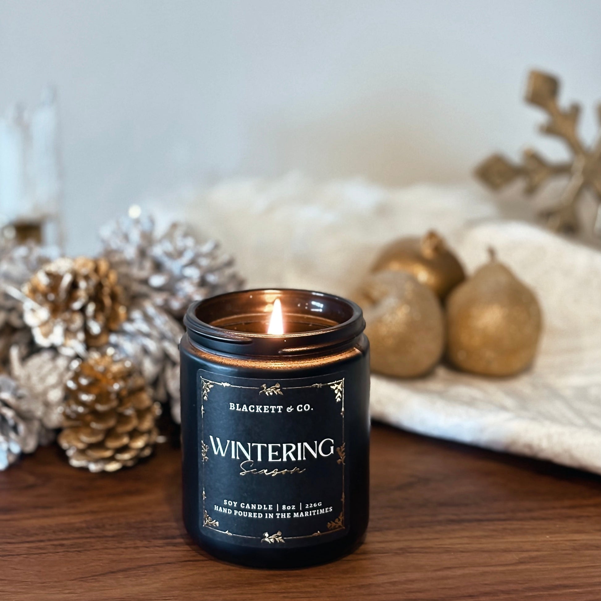 Wintering Season scented natural soy candle by Blackett & Co. on a wooden surface with frosted pine cones and decorative decor in the blurred background.