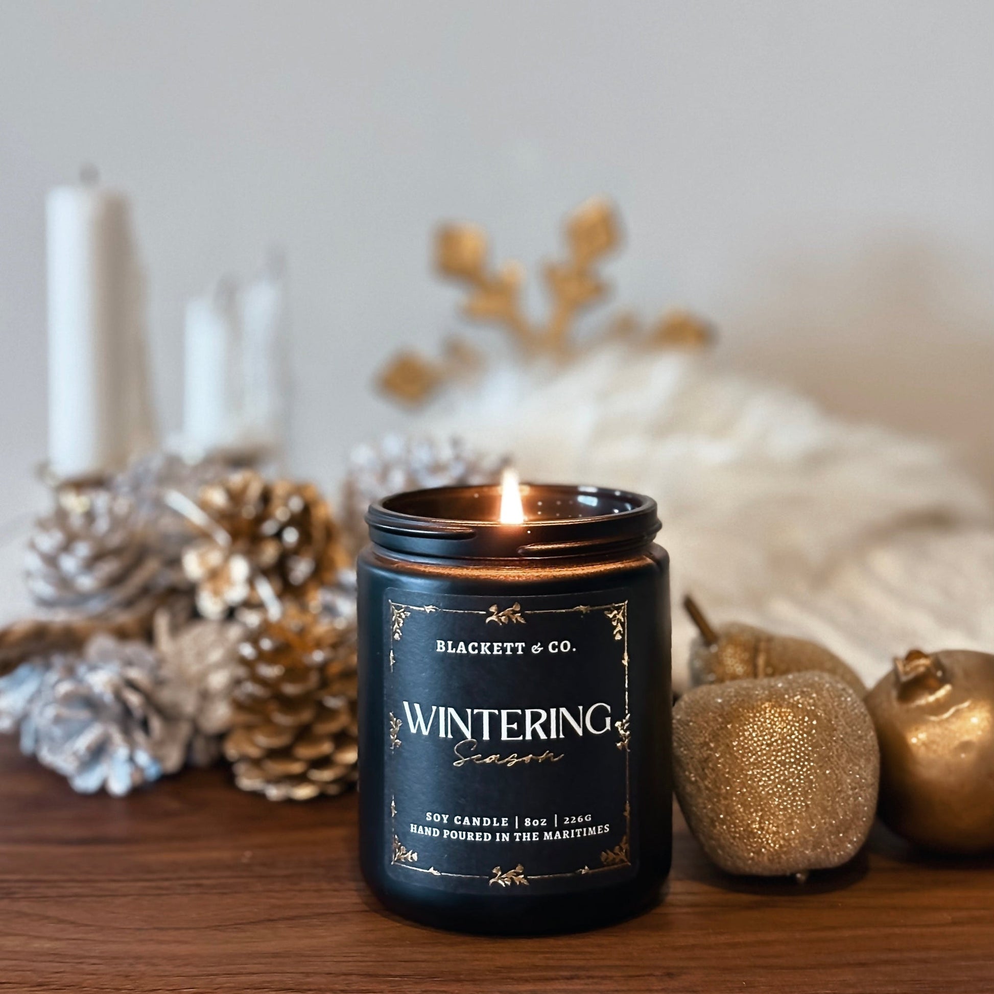 Wintering Season scented natural soy candle on a wooden surface with gold decorative fruit and pine cones.