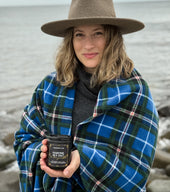 Owner of Blackett & Co. holding a black candle jar of 'Maritime Sea Salt & Honeysuckle' by the ocean, wearing a blue plaid Nova Scotia tartan blanket and brown hat.
