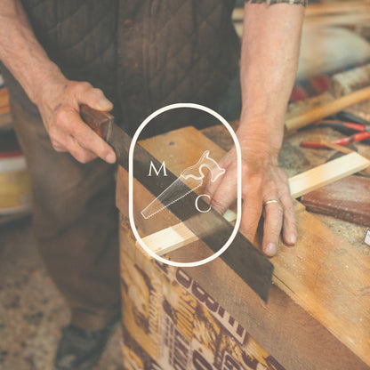 A close-up lifestyle shot of a craftsman using a wooden saw to cut wood on a workbench featuring a prominent white circular watermark overlay in the centre with a hand saw illustration and the letters 'M' and 'C'.