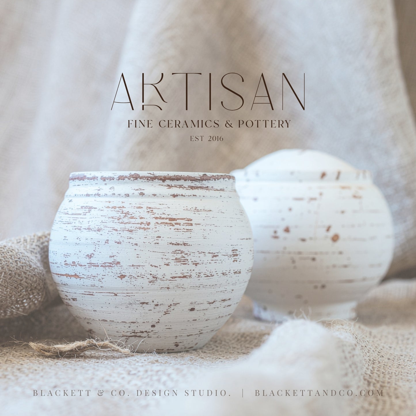 Two small white distressed ceramic vases with a rustic brown speckled finish, displayed on a burlap surface against a soft beige fabric backdrop, with the lettered logo at the top.
