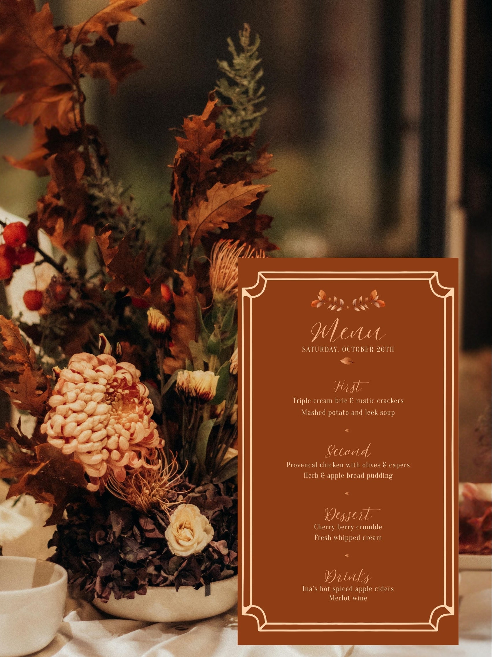 A vertical brown paper menu card with elegant cream typography and decorative corner flourishes, displayed in front of a rustic autumn floral arrangement on a table.
