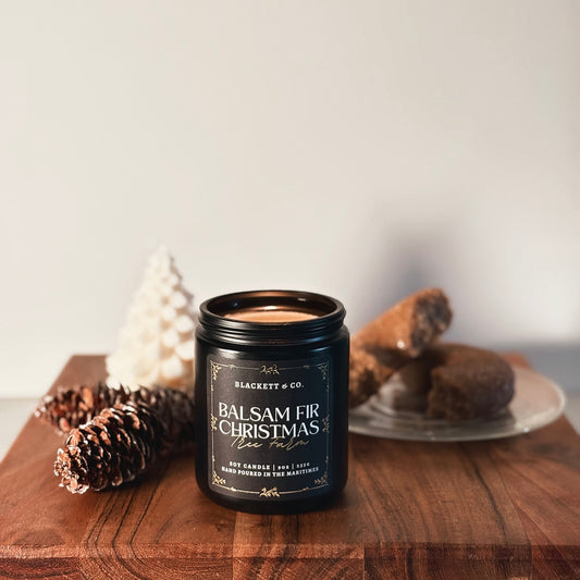 Balsam Fir Christmas Tree Farm scented natural soy candle by Blackett & Co. on a wooden board with a cider doughnut and pine cones.