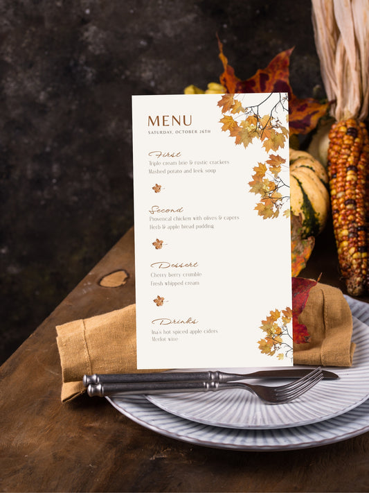 An autumn-themed printed menu card displayed on a plate with a napkin and cutlery, surrounded by fall decor including corn, leaves, and pumpkins on a dark rustic tabletop.