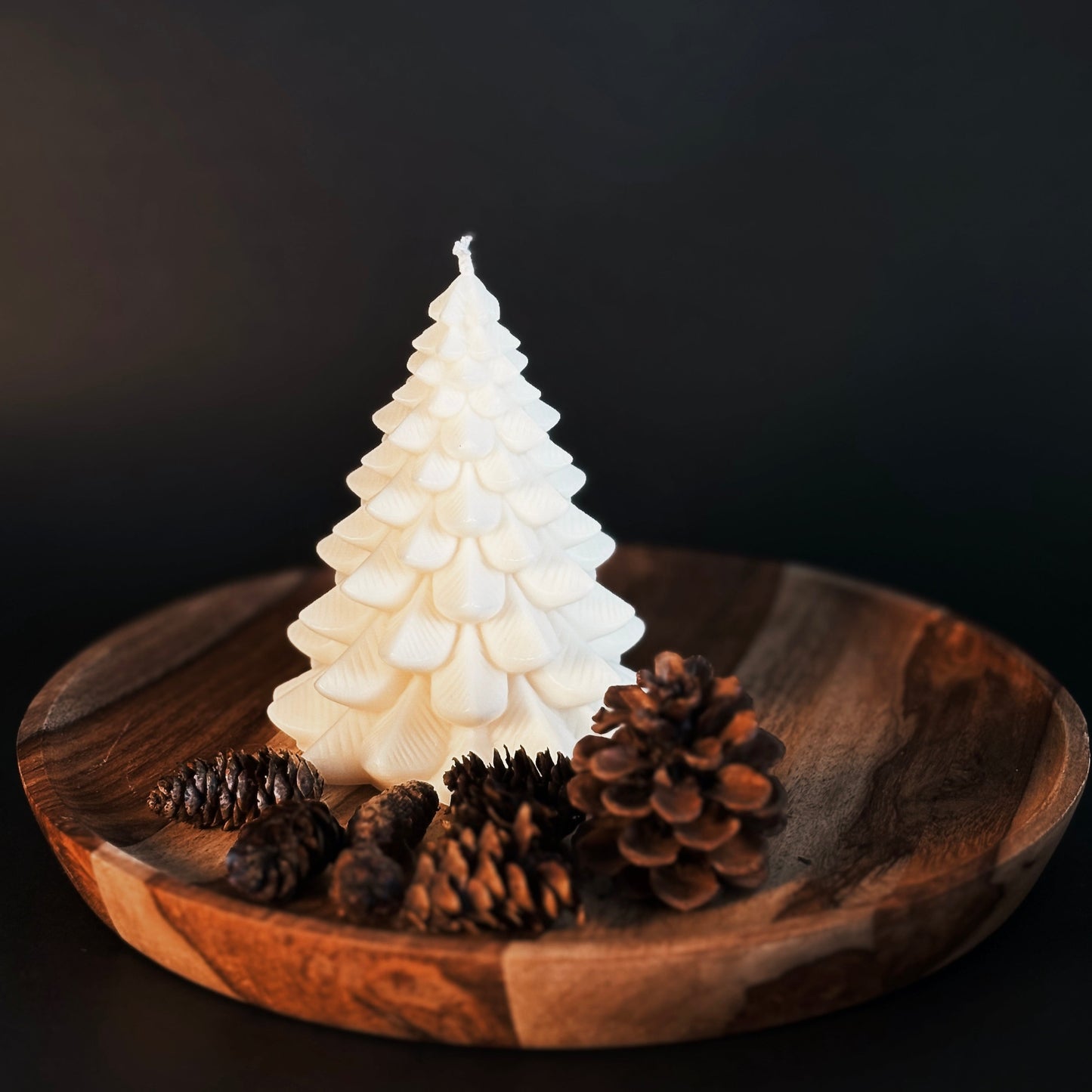 White Christmas tree-shaped soy candle on a wooden plate with pine cones against a black background