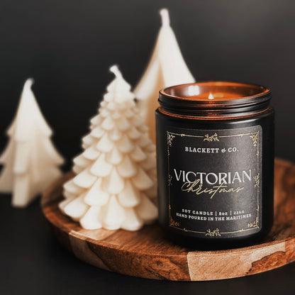 Candle labeled 'Victorian Christmas' by Blackett & Co. on a wooden tray with decorative fir tree-shaped candle in the background.