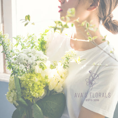 Florals brand logo on an image of florist holding a large bouquet of white and light green hydrangeas, with bright backlighting from a window.