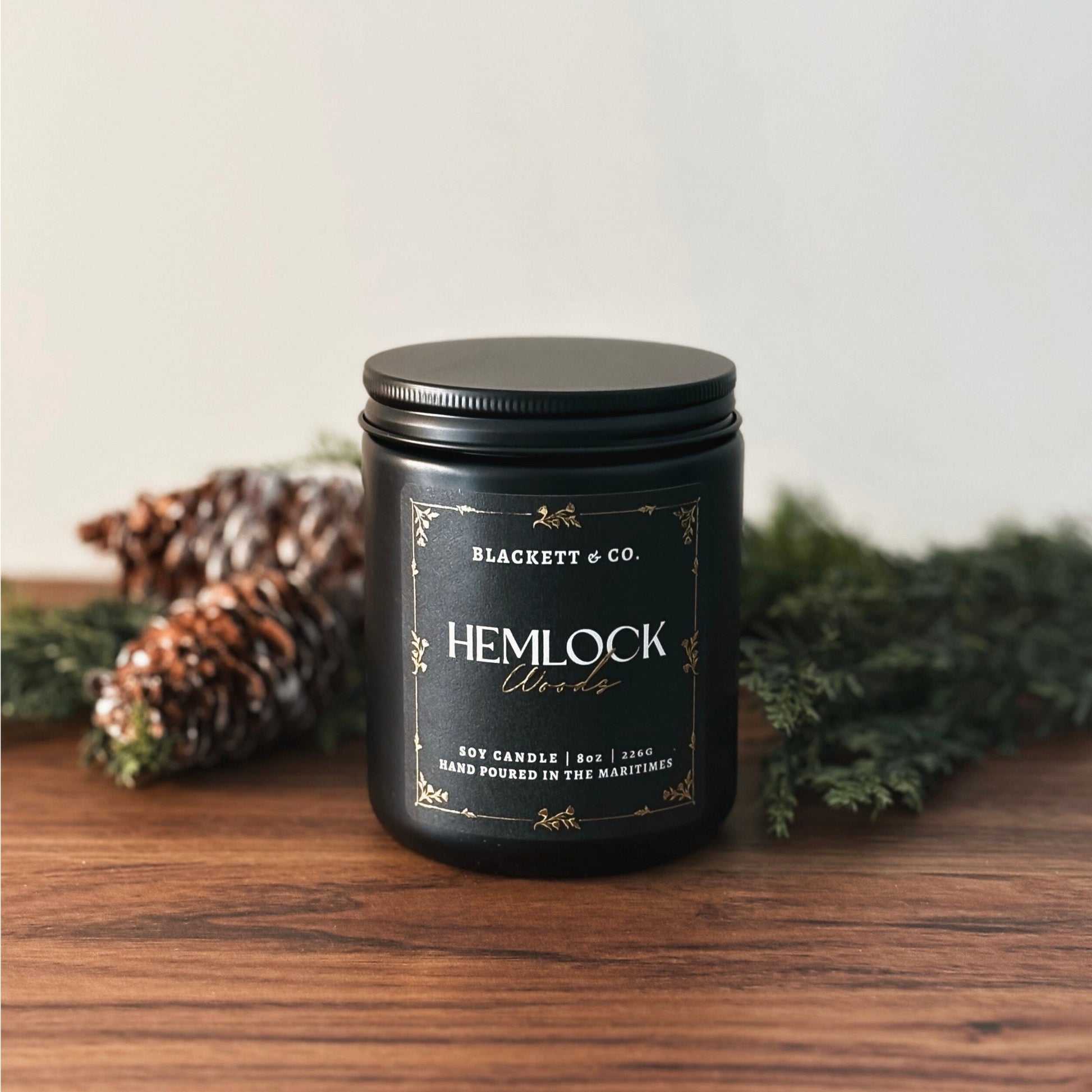 Hemlock Woods scented natural soy candle by Blackett & Co. on a wooden surface with greenery and pine cones.