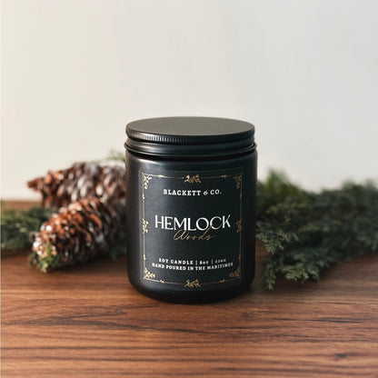 Hemlock Woods scented natural soy candle by Blackett & Co. on a wooden surface with greenery and pine cones.