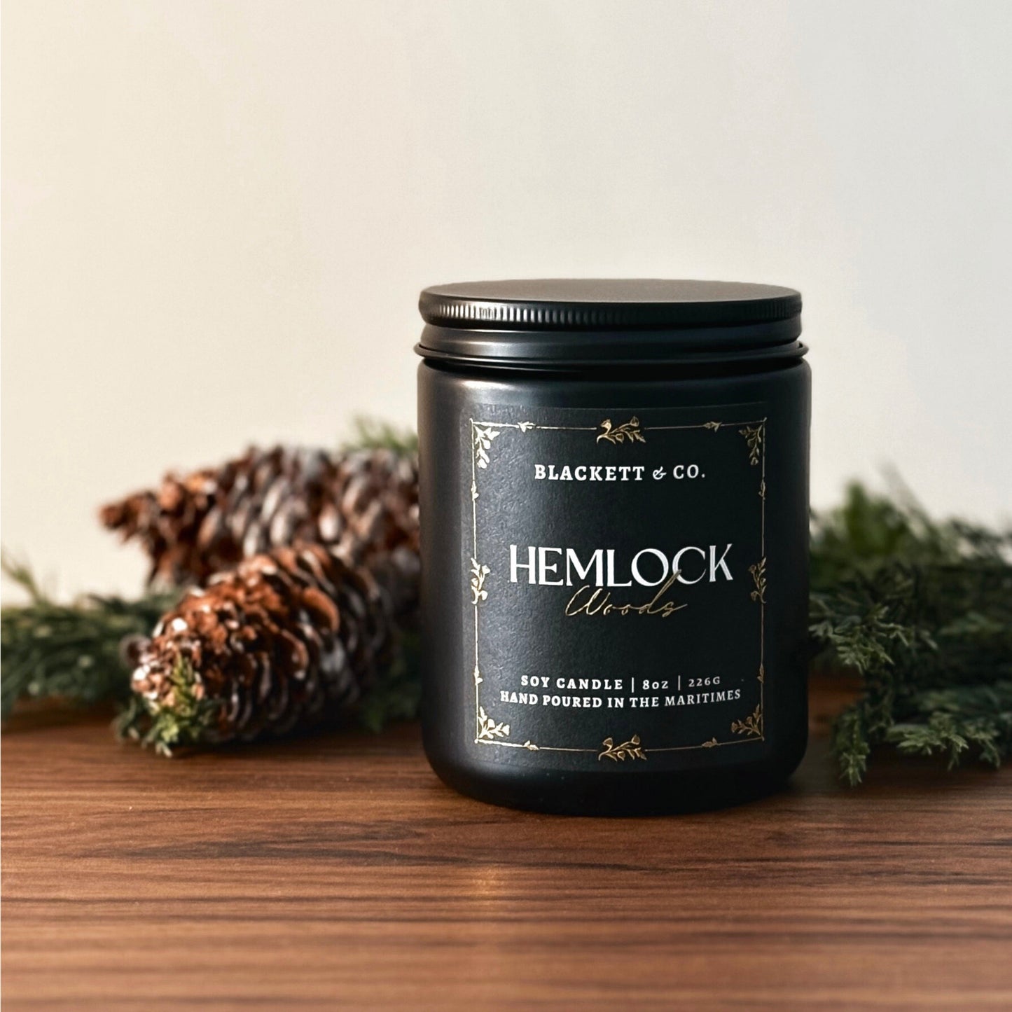 Hemlock Woods scented natural soy candle by Blackett & Co. with pine branches on a wooden surface.