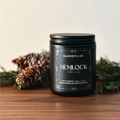 Hemlock Woods scented natural soy candle by Blackett & Co. with pine branches on a wooden surface.