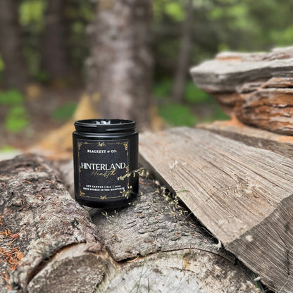 Hinterland Hearth scented soy candle in a black jar by Blackett & Co. sitting on a stack of wooden logs