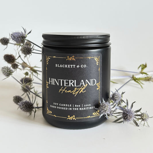Hinterland Hearth scented natural soy candle in a black jar by Blackett & Co. on a light background with thistles