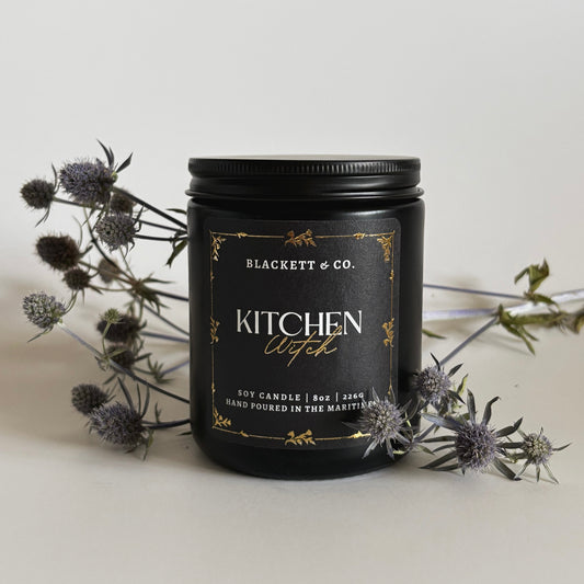 Kitchen Witch scented natural soy candle in a black jar by Blackett & Co. with a light background and thistles