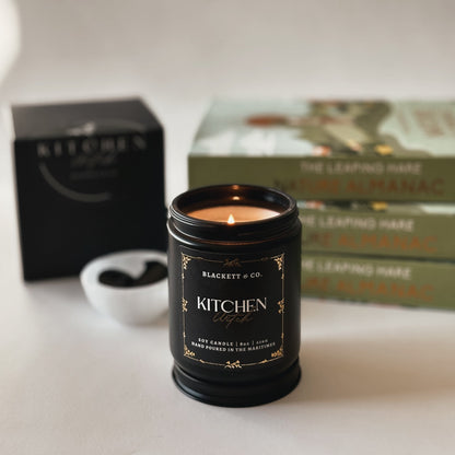 Kitchen Witch scented natural soy candle  handmade by Blackett & Co. on a surface with Nature Almanac books in the background.