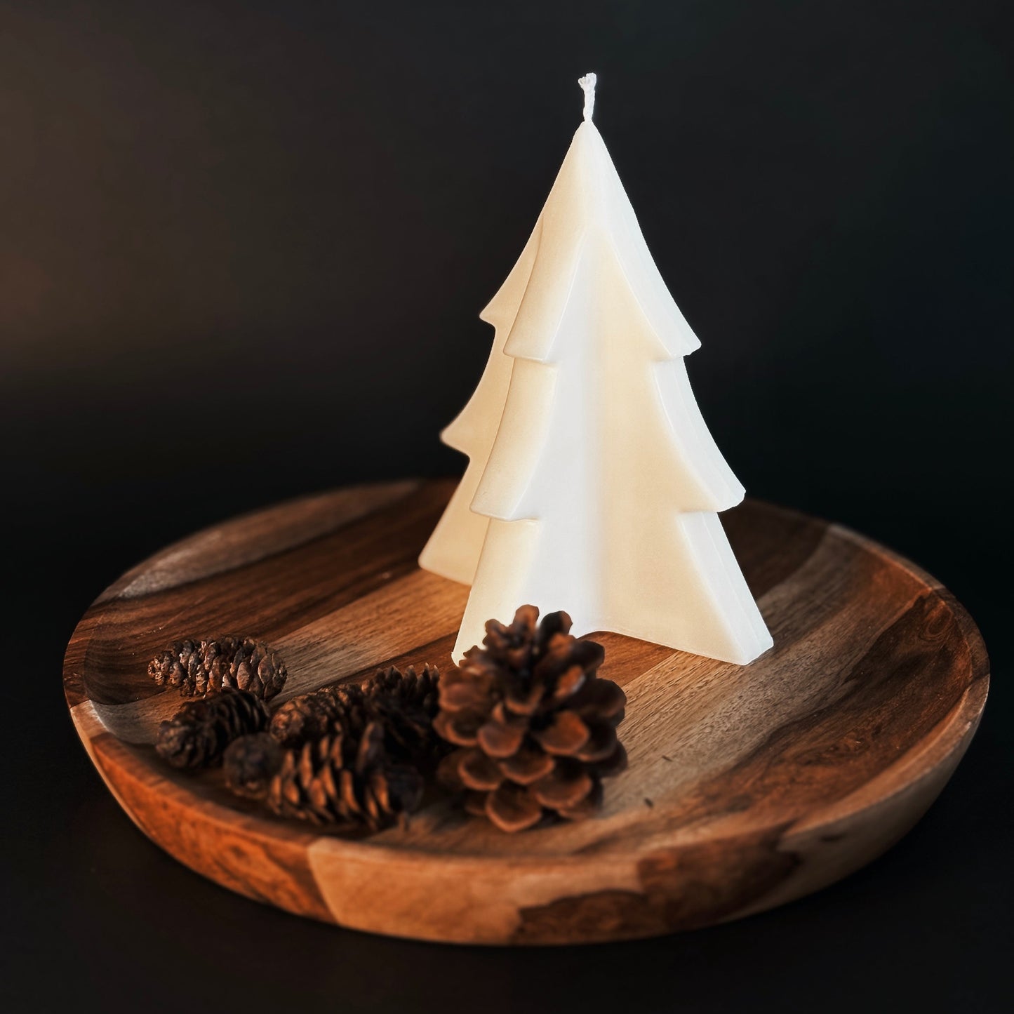 White tree-shaped soy candle on a wooden plate with pine cones against a dark background